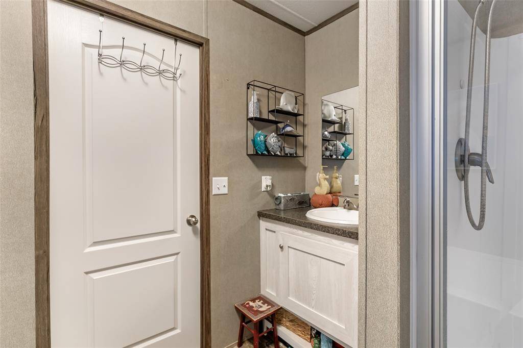 This bathroom features a modern shower, a sink with a countertop, wall-mounted shelves for storage, and a simple door with a towel hook. The neutral tones create a clean and functional space.