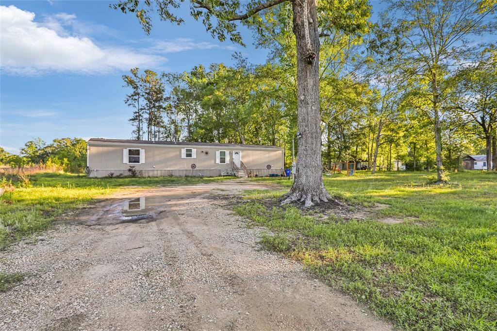 This photo shows a mobile home situated on a spacious, grassy lot with mature trees. A gravel driveway leads up to the home, providing ample outdoor space and a serene, natural setting.