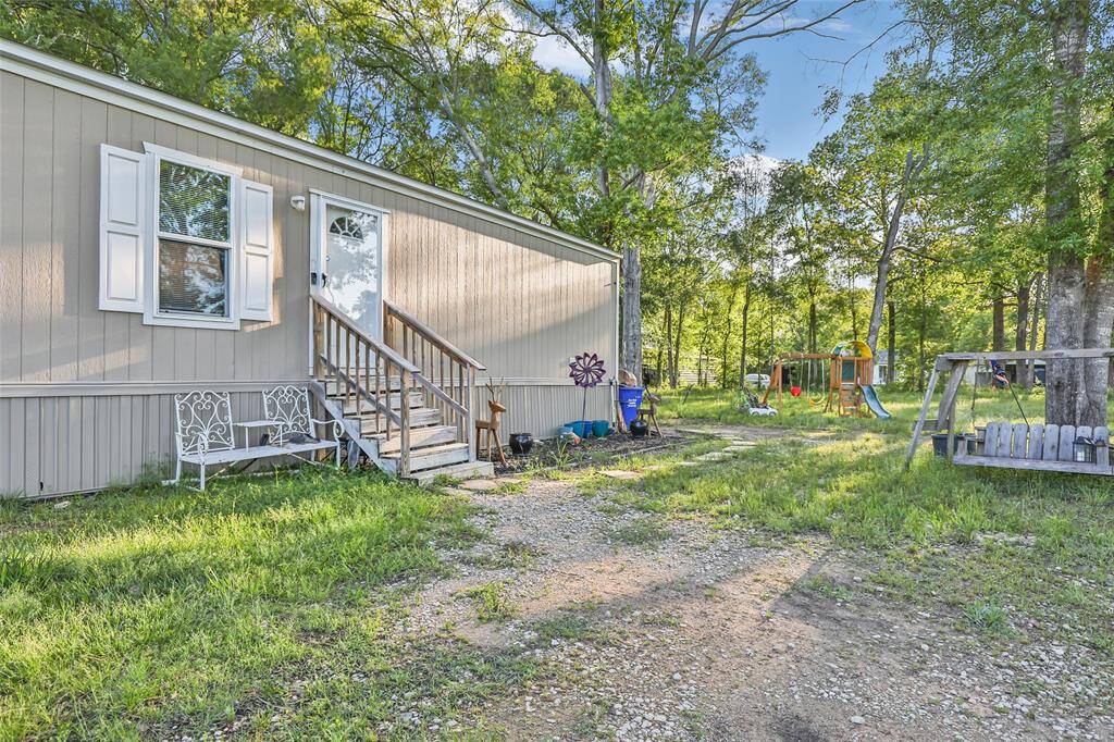This photo shows a cozy mobile home with a small porch and steps leading to the entrance. It's surrounded by a grassy yard with trees, providing a natural, wooded setting. There’s a children’s play area, ideal for families.