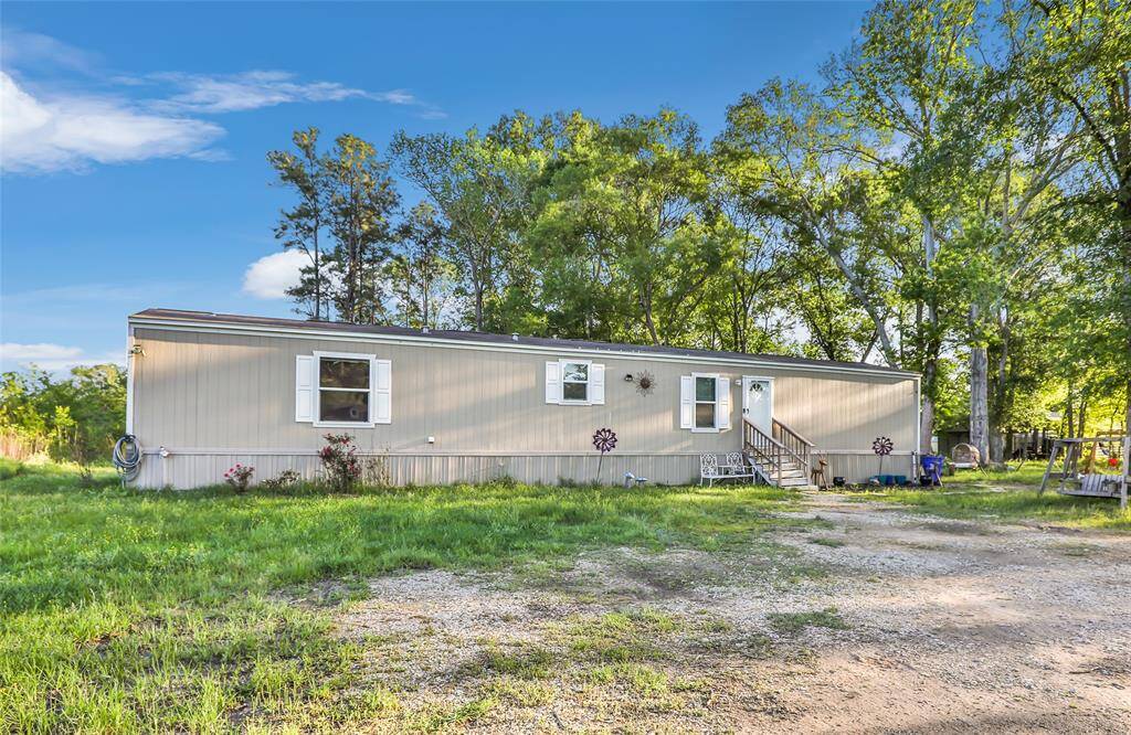 This image shows a single-story mobile home situated on a spacious, grassy lot. The exterior is light-colored with white trim, and there are a few steps leading up to the front door. The surrounding area has mature trees, providing a serene and natural setting.