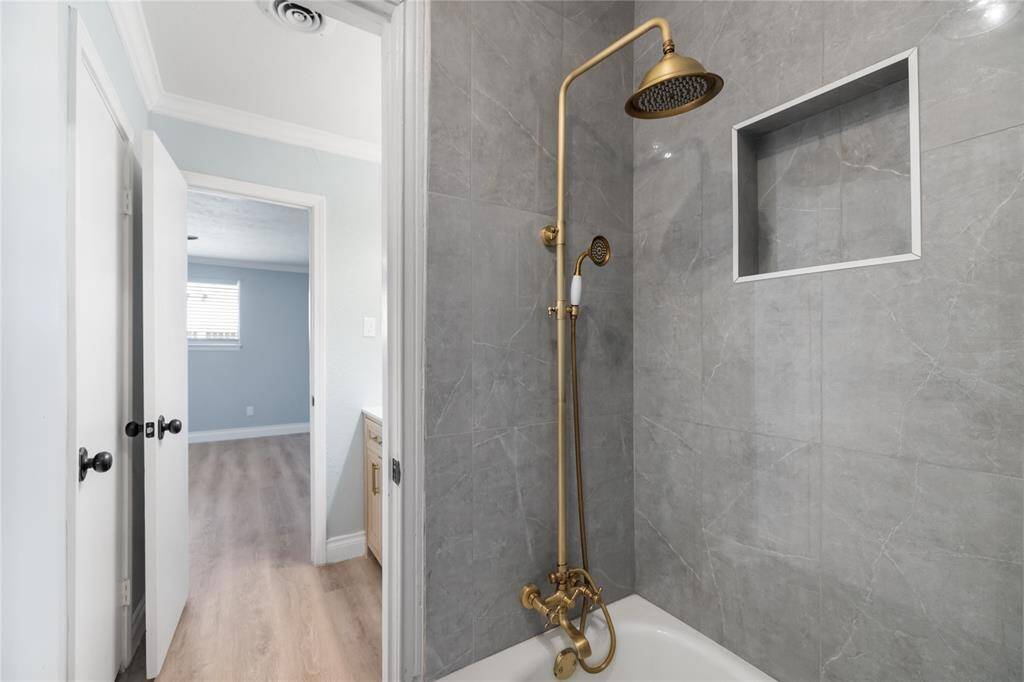 The renovated shower/tub combo features smooth gray stone wall tiles, with a tall rainfall showerhead and a handheld shower option, all accented in elegant gold. The gold-colored faucets add a sophisticated touch, enhancing the modern, luxurious feel of the space.