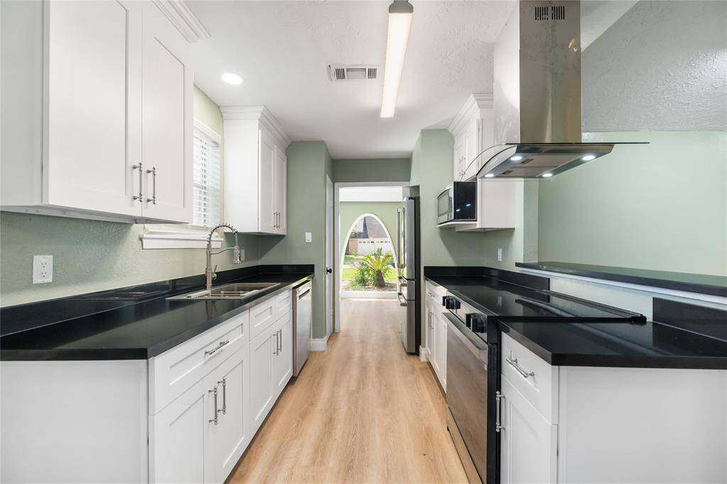 A view from the breakfast area looking back toward the receiving room entrance showcases the spacious, galley-style kitchen layout. Dual countertops on each side provide balance and function, highlighting the white soft-close cabinetry, black countertops, and modern stainless steel finishes. The open design enhances the flow between rooms, perfect for everyday living and entertaining.