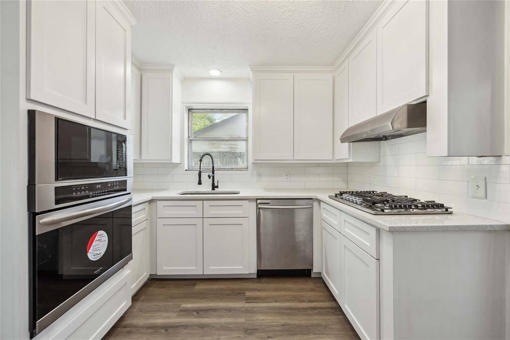 This bright kitchen features modern white cabinetry, stainless steel appliances, a built-in oven, and a gas cooktop. The window above the sink provides natural light, while the wood flooring adds warmth to the space.