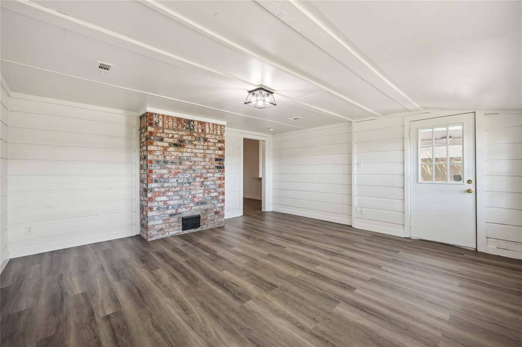Bright, spacious room with wood-style flooring, shiplap walls, and a rustic brick fireplace. It features a modern ceiling light and a door with a window, offering a cozy yet airy feel.