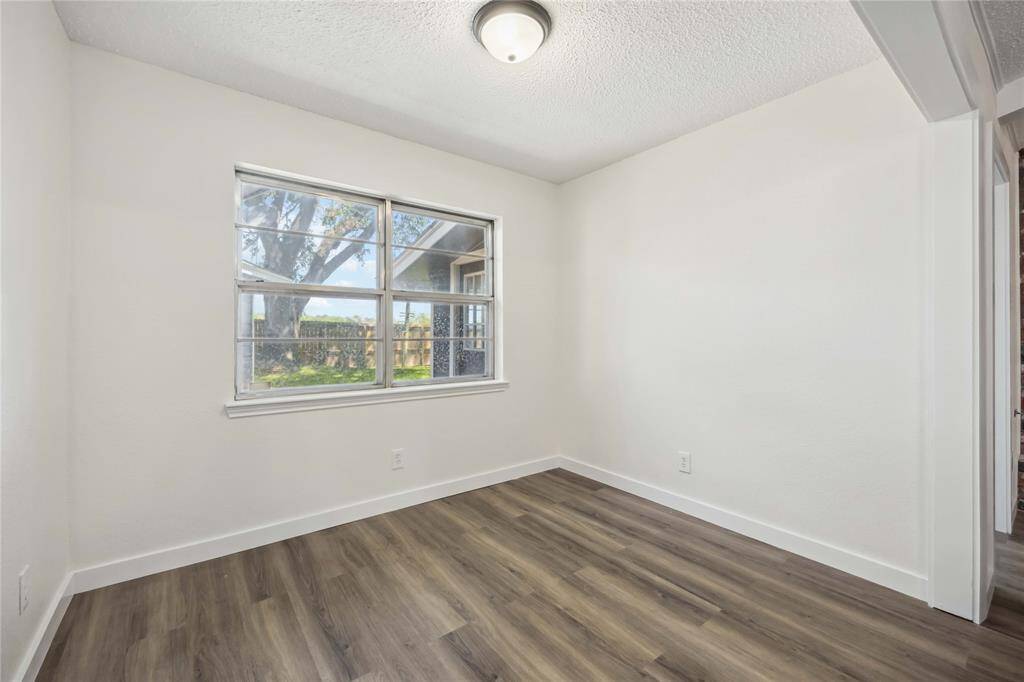 This room features freshly painted white walls and wood-look flooring, creating a clean, modern feel. A large window provides ample natural light and offers a view of the outdoors. Ideal for a cozy home office or small bedroom.