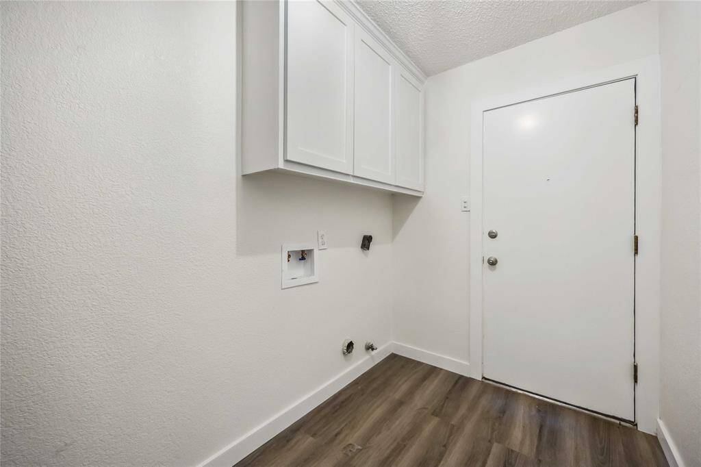 This photo shows a compact laundry room with white walls and cabinets. It features hookups for a washer and dryer, with wood-style flooring and a simple white door.