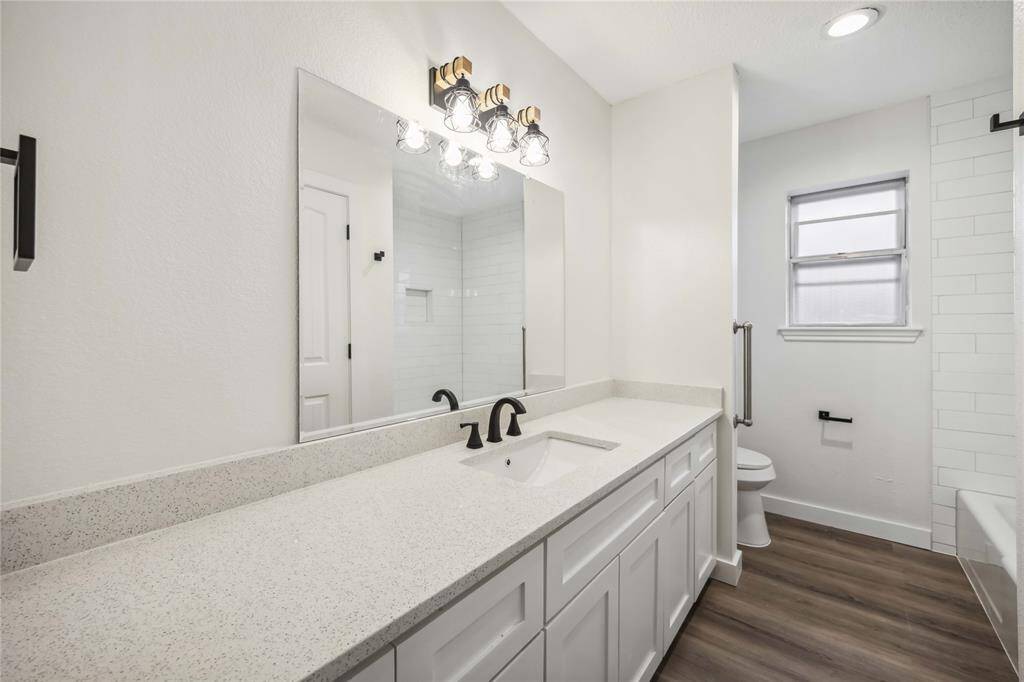 This bathroom features a modern design with a spacious vanity, sleek black fixtures, and a large mirror. There's ample natural light from the window, and the room has a clean, neutral palette with wood-style flooring and subway tile accents.