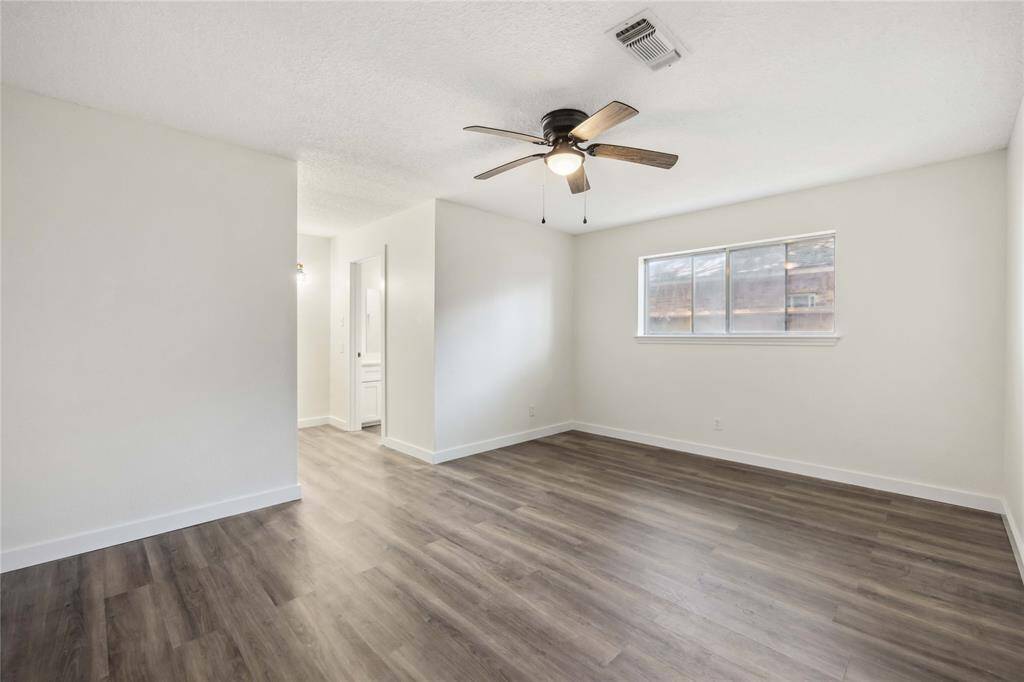 This bright, spacious room features modern laminate flooring, a ceiling fan, and a large window for natural light. The neutral walls offer a versatile backdrop, and there's an open entryway leading to an adjacent area.