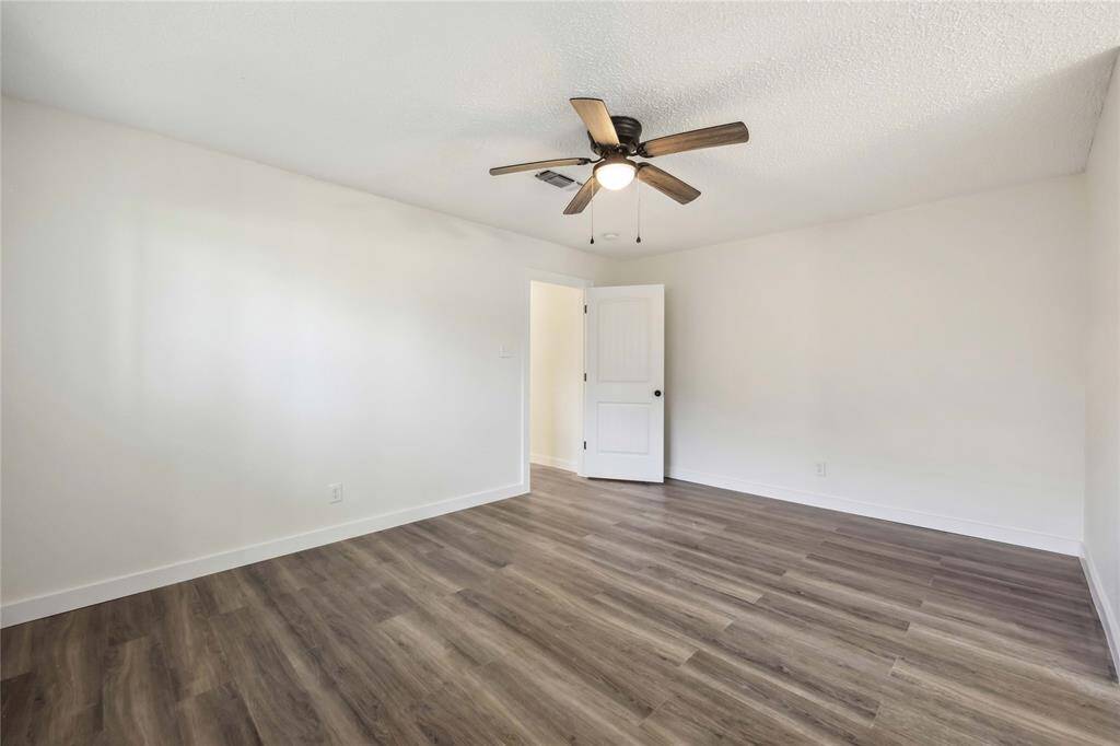Bright, spacious room with light walls and wood flooring, featuring a ceiling fan and an open door leading to another area. Ideal for a bedroom or office.
