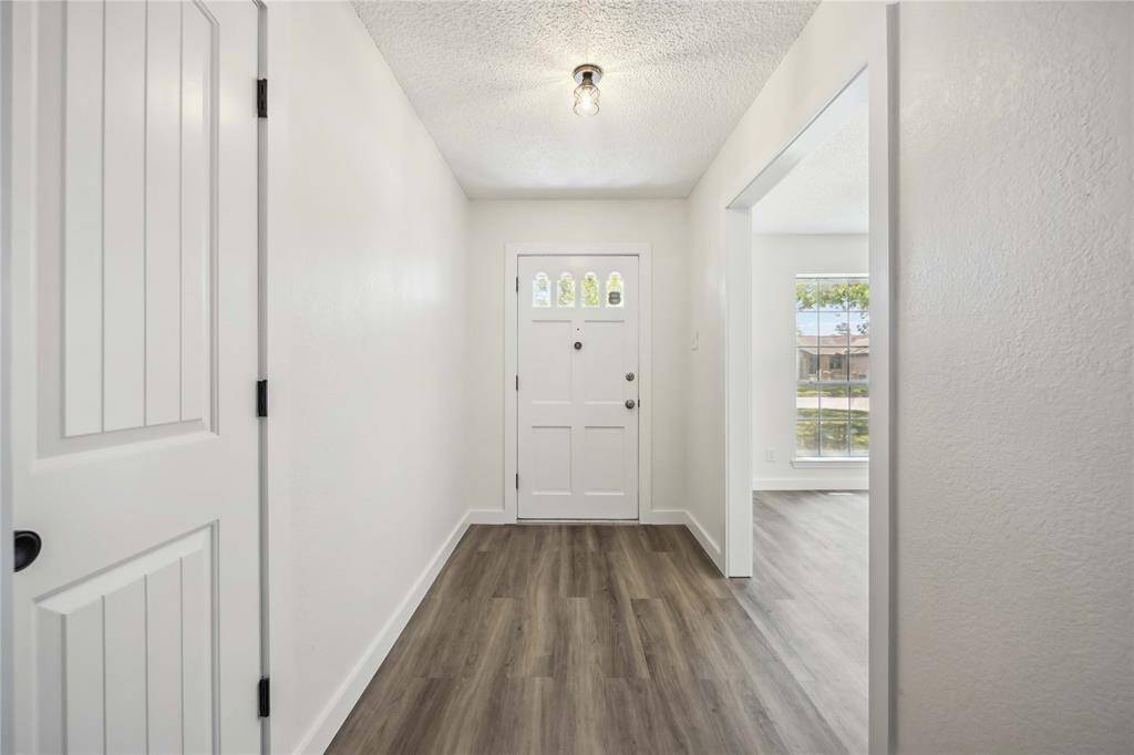 Bright and inviting entryway with light-colored walls and wood-style flooring, featuring a charming front door and a view into a sunlit room with large windows.
