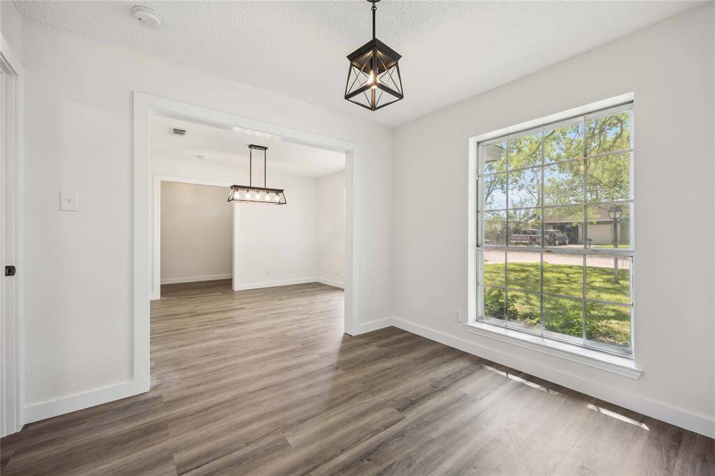 Bright and inviting room with large windows overlooking a green lawn. Features modern light fixtures, light-colored walls, and wood-style flooring, creating a spacious and welcoming atmosphere.