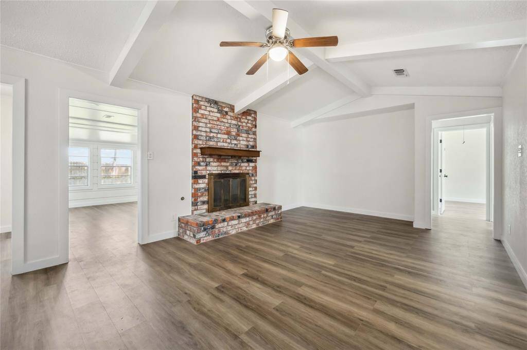 This spacious living room features vaulted ceilings with exposed beams, a rustic brick fireplace, and modern wood flooring. Bright natural light flows in from adjacent rooms, creating a welcoming atmosphere.