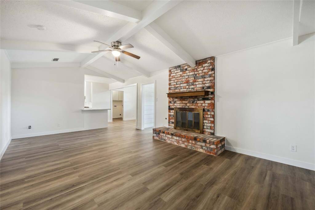This spacious living area features a vaulted ceiling with exposed beams, a ceiling fan, and a rustic brick fireplace. The room has light-colored walls and wood-style flooring, creating a warm and inviting atmosphere. An open layout leads to an adjacent kitchen area.