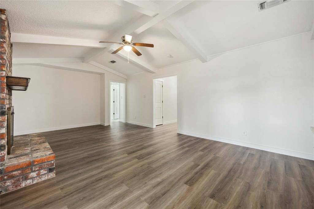 This spacious living room features a vaulted ceiling with exposed beams, a ceiling fan, and a brick fireplace. The room is bright with neutral walls and wood-look flooring, offering a warm and inviting atmosphere.