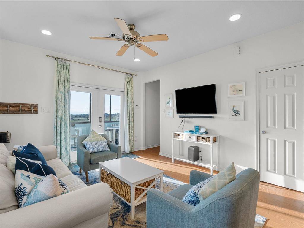 Bright and inviting living room featuring a cozy seating area with a sofa and armchairs, a ceiling fan, and a wall-mounted TV. The room has a modern coastal vibe with ocean-themed decor, light wood look flooring, and French doors leading to a balcony with scenic views.