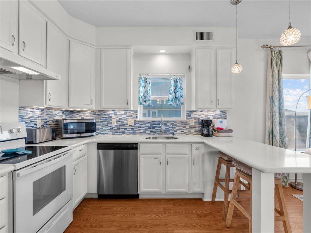 Bright kitchen with white cabinetry and stainless steel appliances. Features a stylish blue mosaic backsplash, hardwood look floors, and a cozy breakfast bar with pendant lighting.