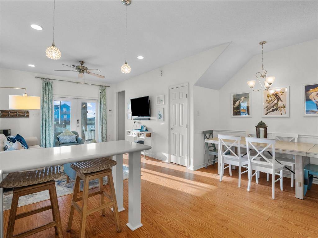 This bright and airy open-concept living space features hardwood look floors, a modern kitchen island with bar seating, and a cozy dining area. Large windows provide natural light and a view of the outdoors, complemented by contemporary lighting and tasteful decor.
