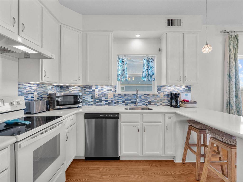 Bright, modern kitchen with white cabinetry and a stylish blue tile backsplash. Features stainless steel appliances, a cozy breakfast bar with seating, and hardwood look floors. Ideal for cooking and entertaining.