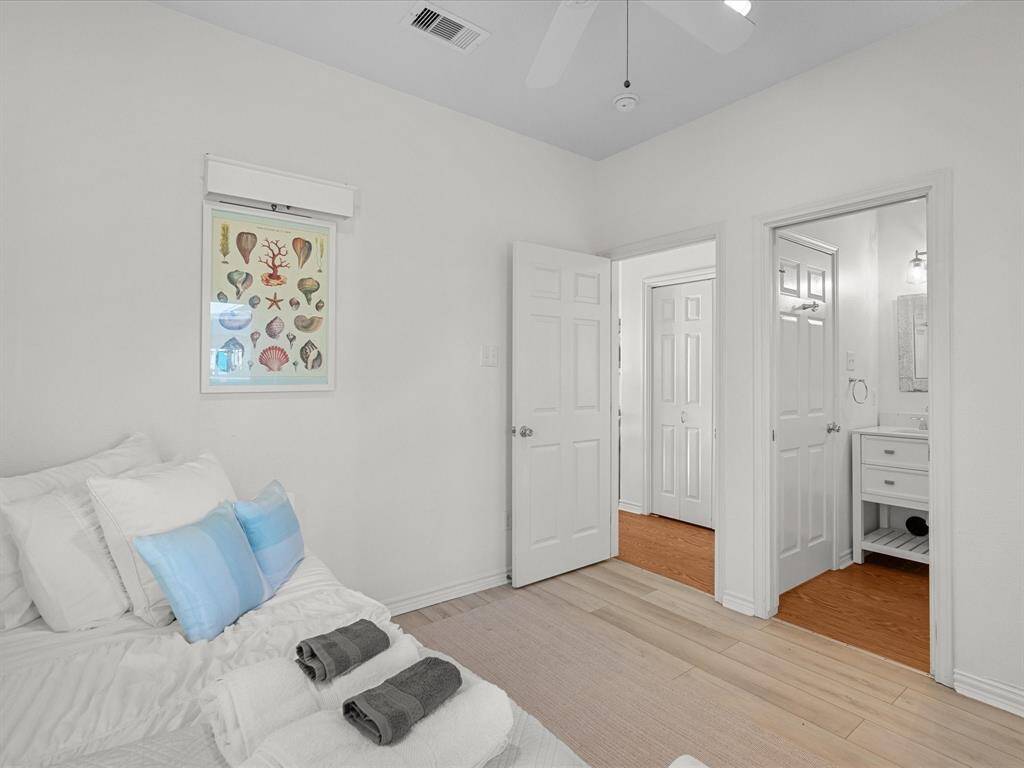 This photo shows a bright, airy bedroom with light-colored walls and flooring. The room features a neatly made bed with white and blue pillows, and towels neatly arranged on it. A ceiling fan is installed overhead, and there's a door leading to an adjacent bathroom with modern fixtures. Artwork decorates the wall, adding a touch of color.