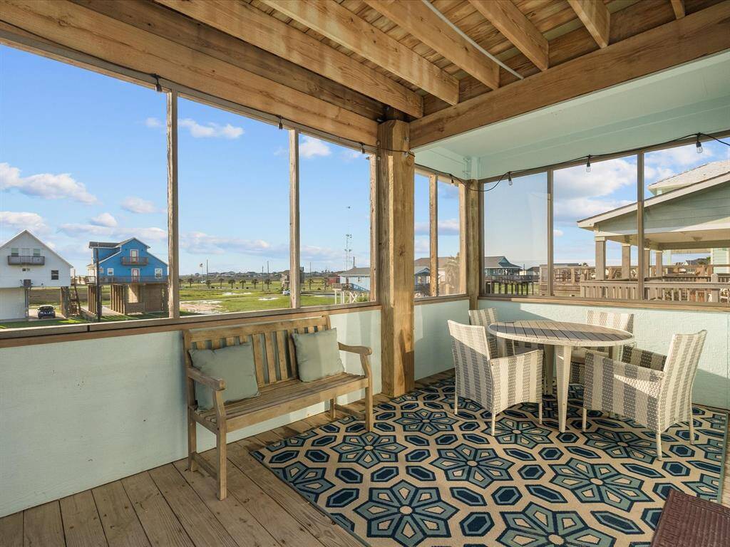 This photo showcases a cozy screened porch with wooden beams and a view of a scenic neighborhood. It features a charming seating area with a wooden bench and a wicker dining set on a patterned rug, perfect for relaxing or entertaining.