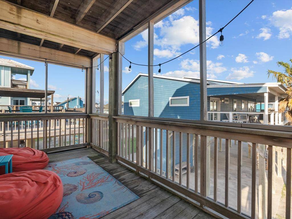This image shows a cozy, screened-in porch with wooden railings and a beamed ceiling. It features comfortable seating with beanbags and a decorative rug. String lights add a charming touch, and the view overlooks neighboring houses under a bright blue sky. Perfect for relaxing or entertaining.