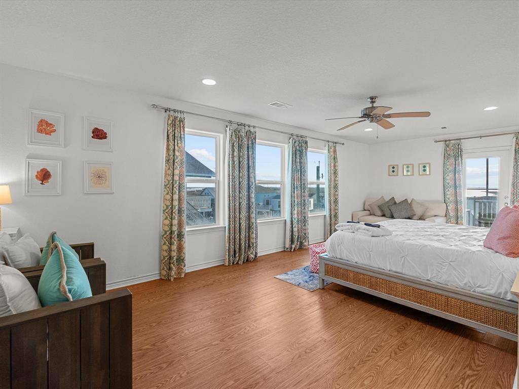 Spacious, bright bedroom with hardwood look floors, ceiling fan, and large windows offering scenic views. Decor includes coastal-themed artwork and patterned curtains.