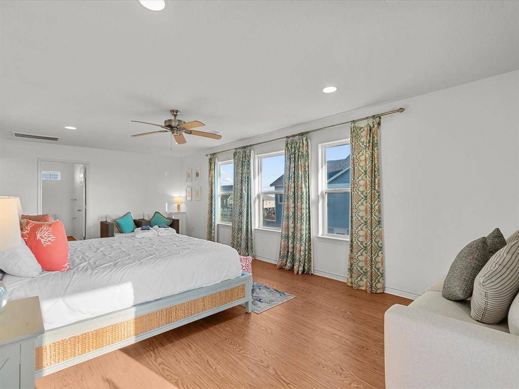 Spacious and bright bedroom with wood look flooring, large windows, and elegant drapes. Features a ceiling fan, cozy seating area, and neutral decor for a relaxing atmosphere.