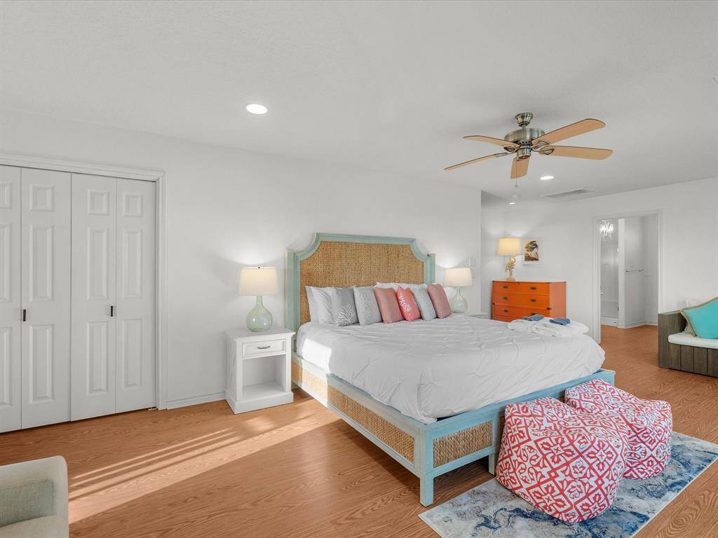 A spacious, bright bedroom with wood look flooring features a large bed with colorful pillows, dual nightstands, a ceiling fan, and ample closet space. The room includes a stylish dresser, cushioned seating, and access to an en-suite bathroom. (see exemptions to non-realty)