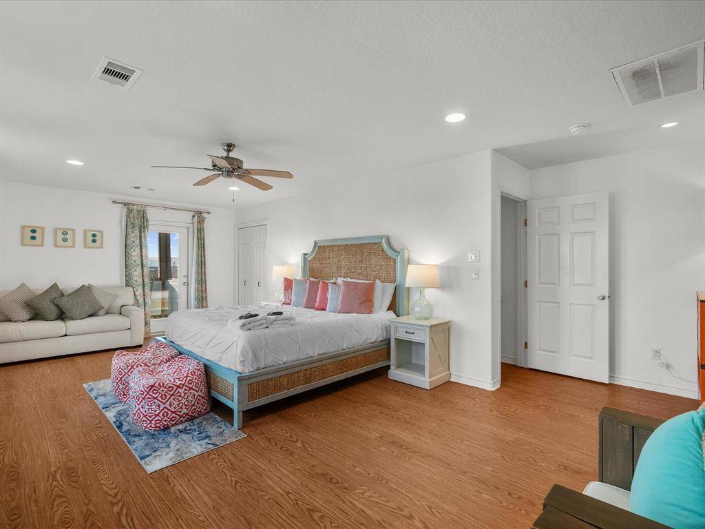 Bright and spacious bedroom featuring a large bed with colorful pillows, hardwood flooring, and a ceiling fan. There's a cozy sofa, decorative poufs, and a door leading to a balcony. The room is well-lit with recessed lighting and has a calming, neutral color scheme.