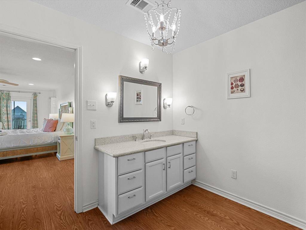 This photo shows a bright bathroom with a white vanity and countertop, featuring a stylish chandelier and wall sconces. The room connects to a cozy bedroom with wood flooring and natural light.
