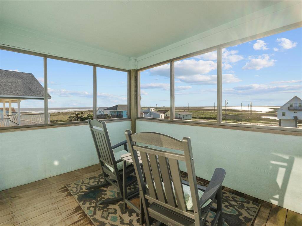 This photo shows a charming, enclosed porch with two rocking chairs and a rug, offering stunning views of a coastal landscape and a bright blue sky. Perfect for relaxing and enjoying the serene surroundings.