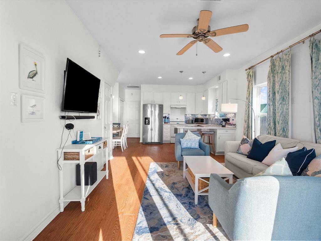 Bright and inviting open-concept living area with hardwood look floors, featuring a cozy seating arrangement, modern kitchen with stainless steel appliances, and a mounted TV. The space is accented by stylish decor, a ceiling fan, and large windows with curtains that allow natural light to fill the room.