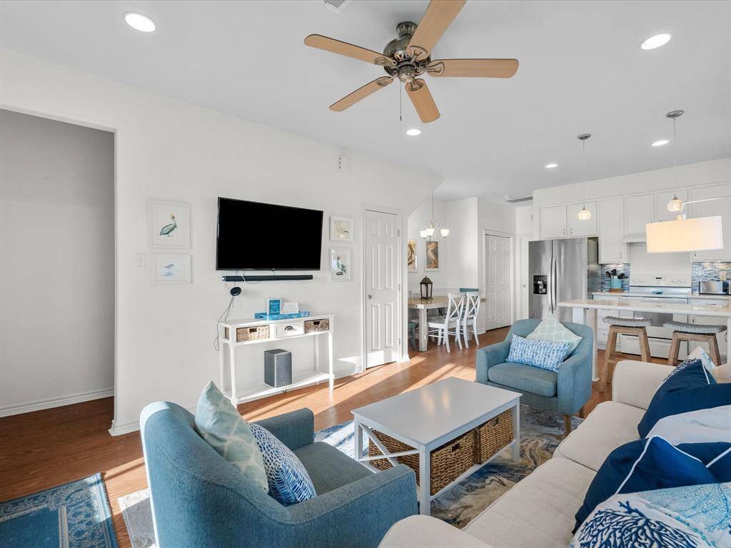 This inviting open-concept living space features a cozy sitting area with a wall-mounted TV, a stylish kitchen with modern appliances, and a dining area. The light color palette, ceiling fan, and recessed lighting create a bright and airy atmosphere, perfect for relaxation and entertaining.