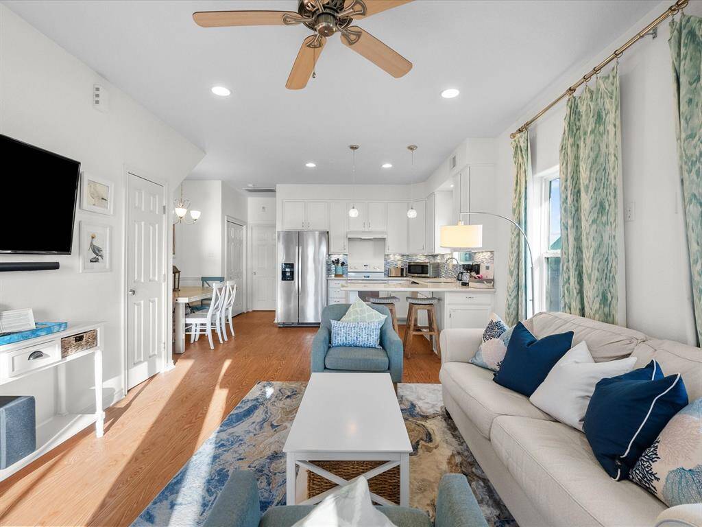 Bright and inviting open-plan living area with hardwood look floors, modern kitchen, and cozy seating. Features include a ceiling fan, stylish decor, and ample natural light. Perfect for entertaining or relaxing.