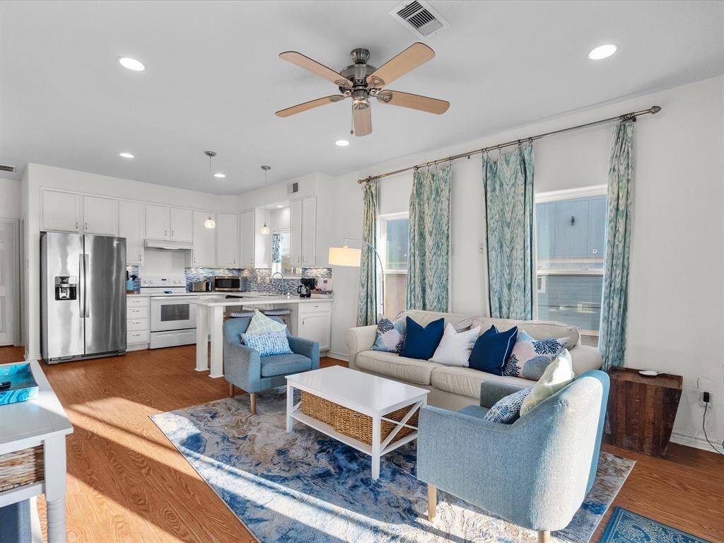 Bright and open living area with wood look floors, featuring a modern kitchen with stainless steel appliances. The space is tastefully decorated with blue accents, a ceiling fan, and natural light from multiple windows.