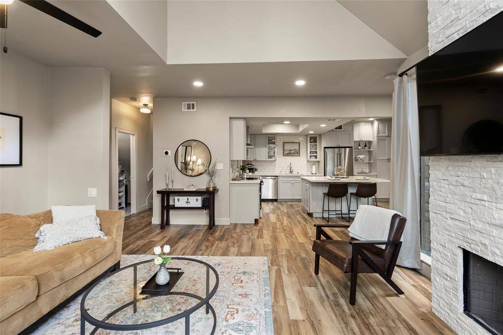 This inviting living space features a modern kitchen with sleek cabinetry and a spacious island, seamlessly flowing into a cozy living area. The room has warm wood flooring, a stylish fireplace, and ample natural light, creating a welcoming atmosphere perfect for relaxation and entertaining.
