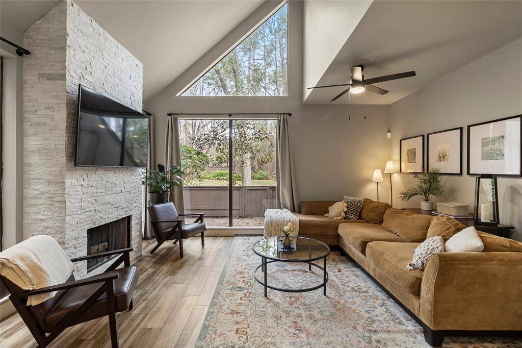 This inviting living room features high vaulted ceilings and large windows that fill the space with natural light. A modern stacked-stone fireplace serves as a striking focal point, while views of the outdoor greenery enhance its welcoming atmosphere.