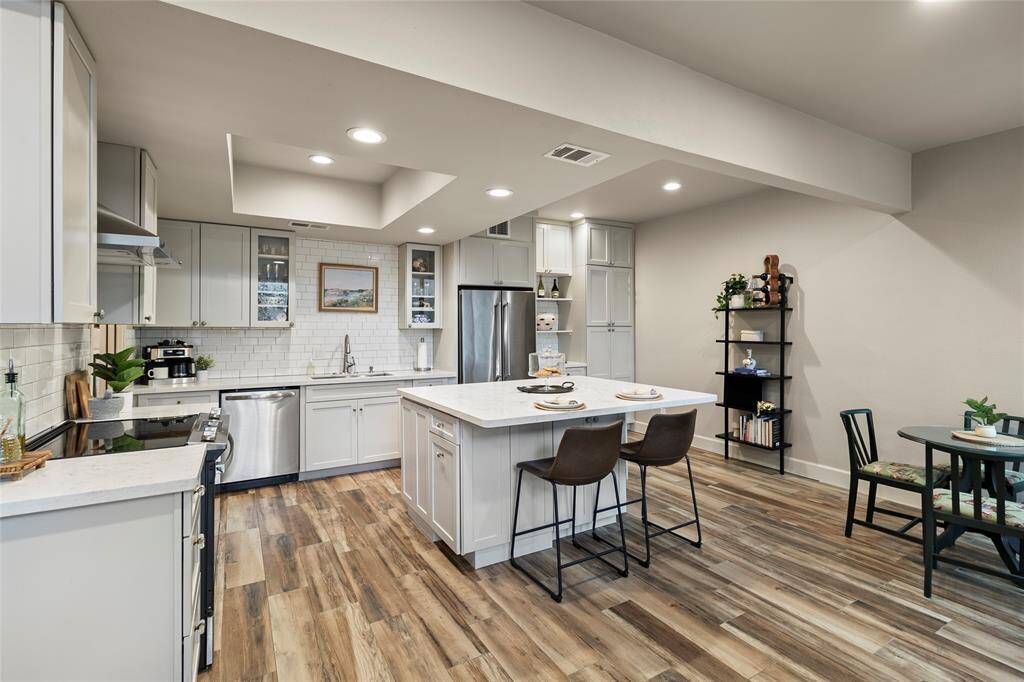 A modern, open-concept kitchen with an eat-in island, stainless steel appliances, and light cabinetry. The kitchen seamlessly connects to a cozy living area, featuring wood-look flooring and recessed lighting.