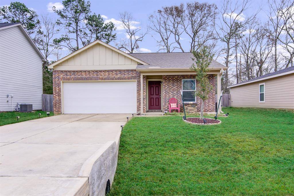 This charming single-story home features a brick facade and a two-car garage. The front yard is well-maintained with a small tree and a cozy porch with seating. It's situated in a quiet neighborhood with lush greenery and mature trees in the background.