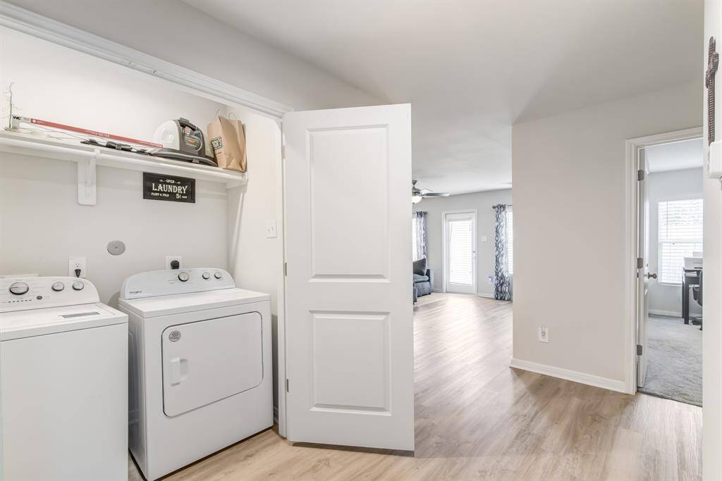 This photo shows a bright, open laundry area with a washer and dryer, adjacent to a living room with natural light and wood like flooring. The layout offers seamless flow between spaces, enhancing functionality.