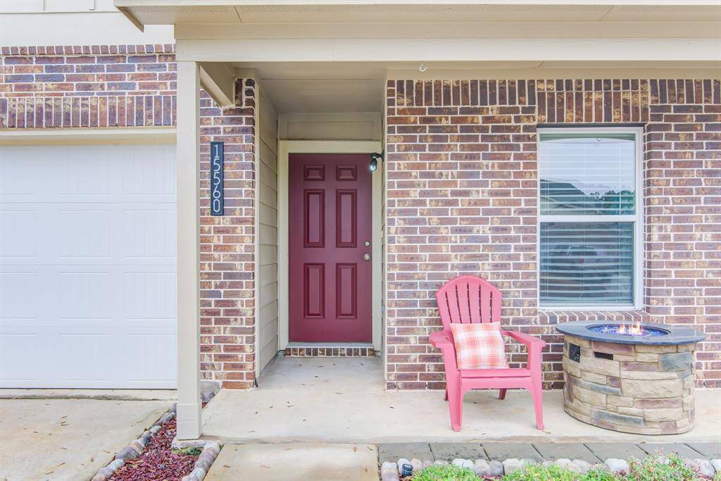 Charming front porch with a brick facade features a welcoming red door. There's a cozy seating area with a pink chair and a small fire pit, perfect for relaxing. A large window and neatly landscaped edge complete the inviting entrance.
