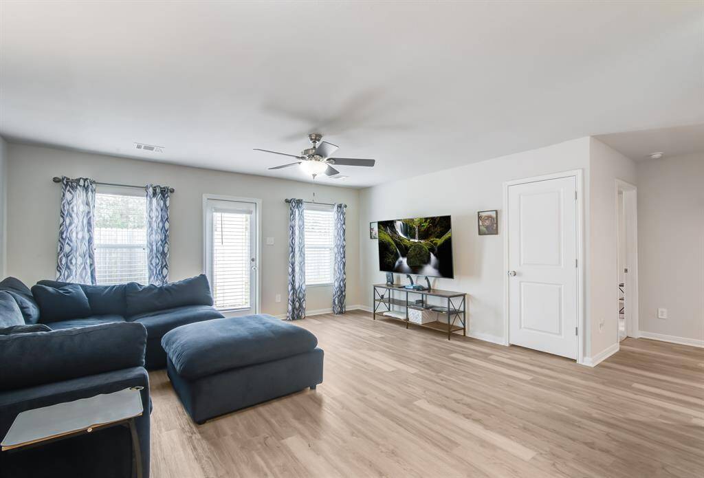 Bright and spacious living room with like wood flooring, a large navy sectional sofa fits perfectly, and a wall-mounted TV. It features abundant natural light from the windows and a door leading outside, complemented by modern curtains and a ceiling fan.