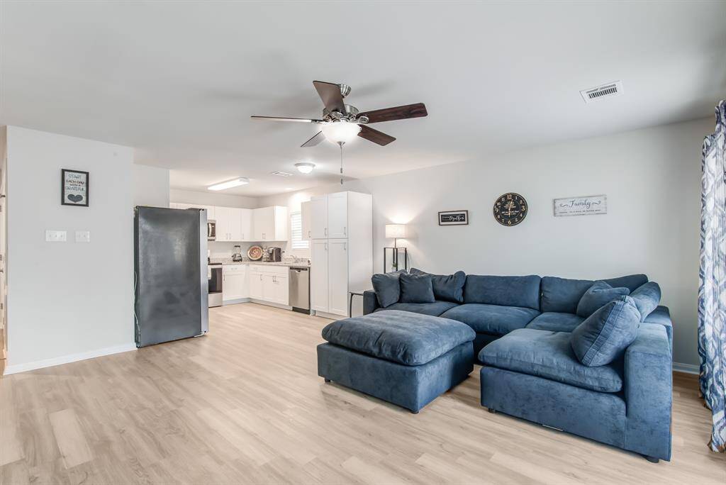 This open concept living space features a cozy blue sectional, like wood flooring, and a modern kitchen with white cabinets and stainless steel appliances. A ceiling fan and natural light add comfort and brightness.