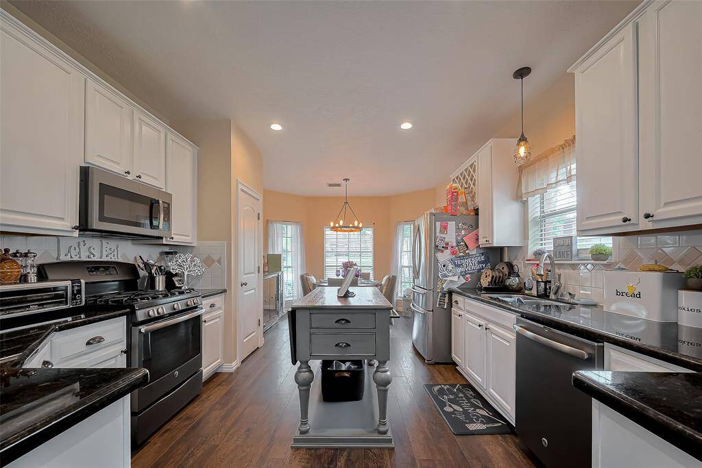 The kitchen features modern stainless steel appliances, white cabinetry, and dark countertops. It has a central island, hardwood floors, and ample natural light from the adjacent dining area.