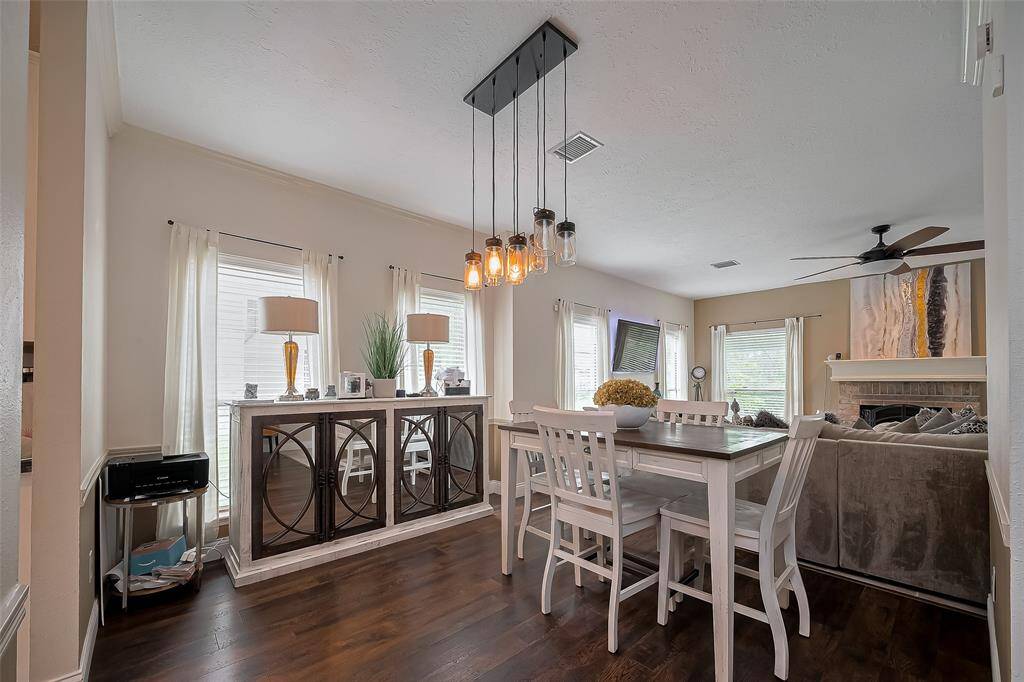 Bright and inviting dining area with hardwood floors, modern pendant lighting, and large windows. The space flows into a cozy living room featuring a ceiling fan and fireplace, creating a perfect setting for entertaining or relaxing.