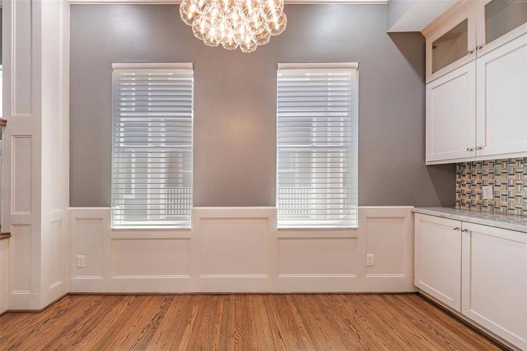 Bright dining area with elegant wainscoting, hardwood floors, and a modern chandelier. Features large windows with blinds and built-in cabinetry with a tiled backsplash for added storage and style.