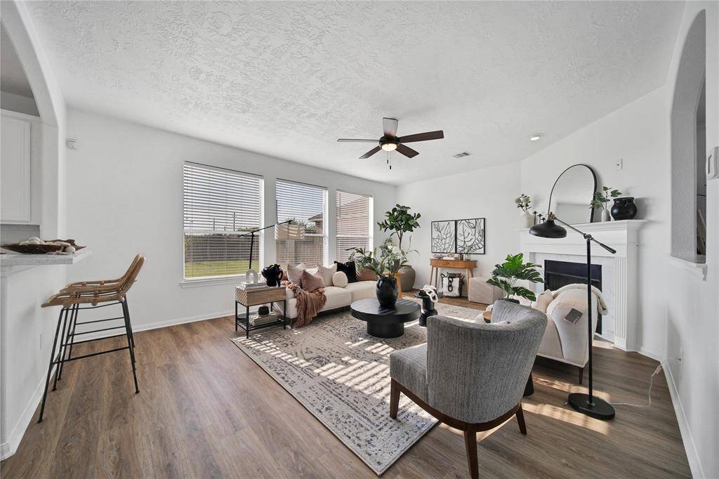 This inviting living room features a modern design with light walls and wood-like vinyl plank flooring. It includes a cozy fireplace, large windows for ample natural light, and a ceiling fan. The open layout connects seamlessly to the kitchen, perfect for entertaining.