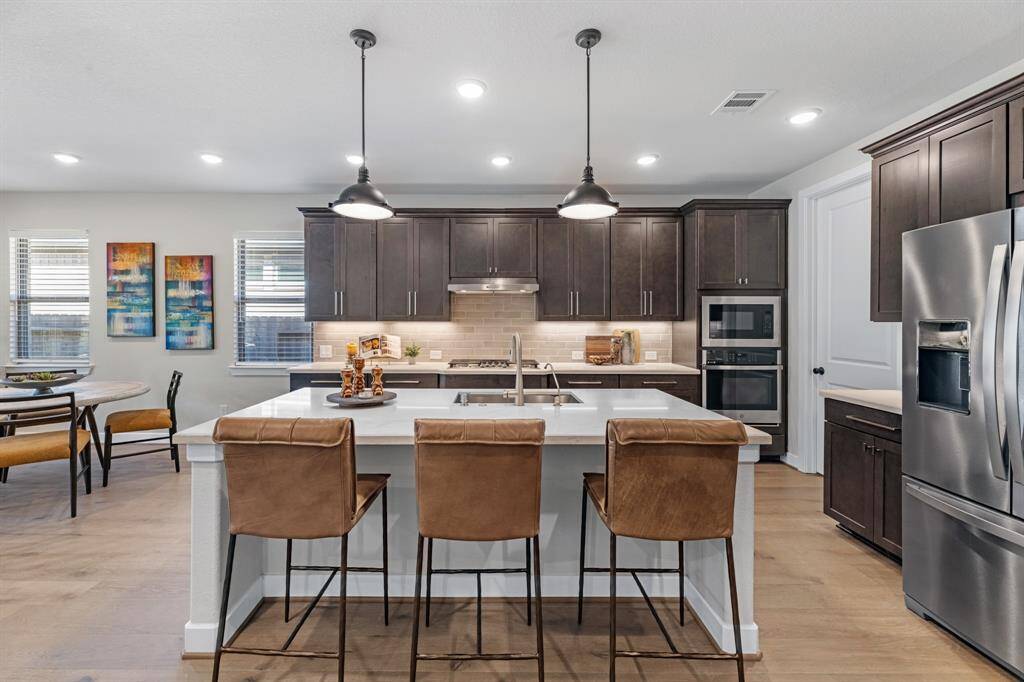 This open-concept kitchen features dark wood cabinetry, stainless steel appliances, and a central island with seating. The space is bright with large windows, warm wood flooring and connection to both the living room and breakfast area