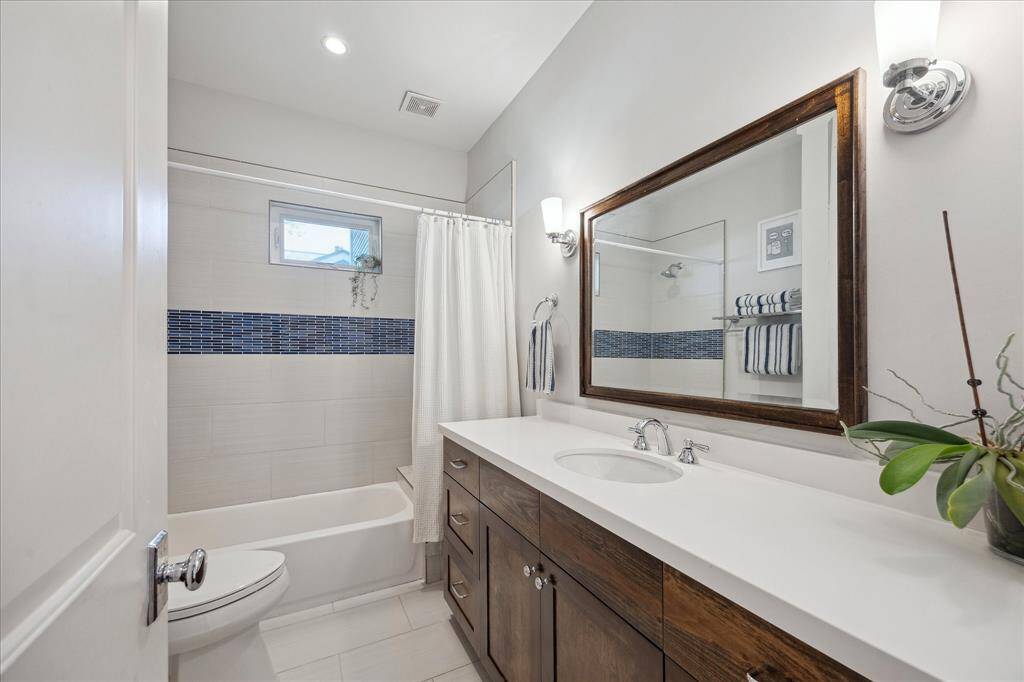 This secondary bathroom features a sleek combination of modern and warm finishes, including a rich wood vanity, crisp white countertops, and a tiled tub/shower with a striking blue mosaic accent band.