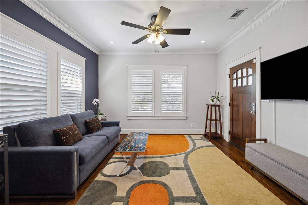 The front living room welcomes you with warm hardwood floors, a sleek ceiling fan, and natural light streaming through classic plantation shutters — creating a comfortable and inviting first impression.